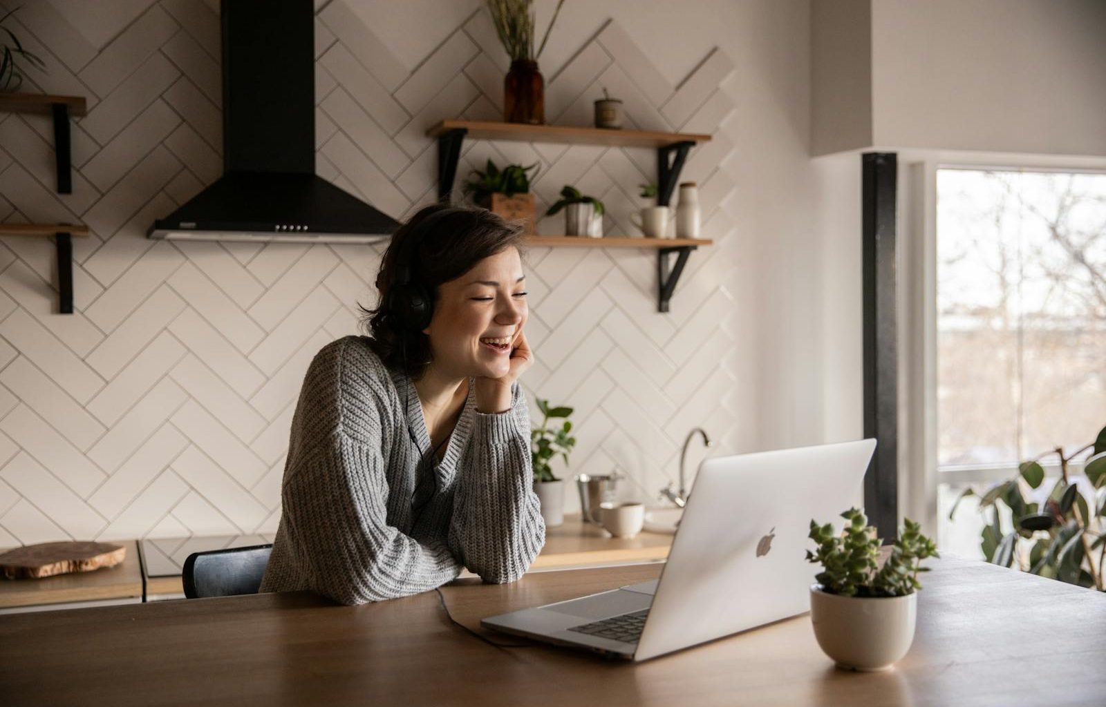 Young cheerful female smiling and talking via laptop while sitting at wooden table in cozy kitchen