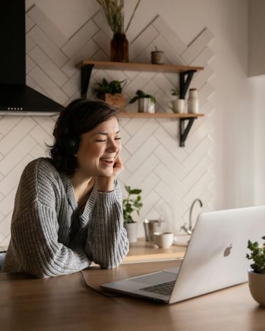 Young cheerful female smiling and talking via laptop while sitting at wooden table in cozy kitchen